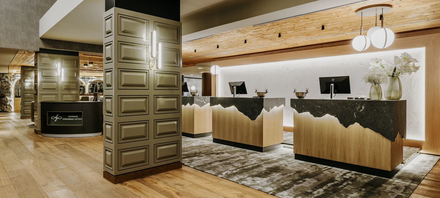 Dieses Foto zeigt den luxuriösen Innenraum des Grand Tirolia Kitzbühl Hotels. Man kann eine luxuriöse Lobby mit einer modernen Rezeption, bequemen Sitzmöbeln, einem hübschen Blumenarrangement und einer riesigen Glasfront erkennen.